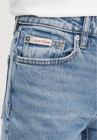 Jeans in denim azzurro chiaro con design aderente, caratterizzati da un'etichetta Calvin Klein rossa e bianca sulla tasca posteriore e dettagli in metallo argentato.