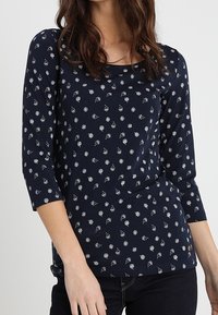 Tummansininen pitkähihainen paita, jossa on pyöreä pääntie. Paidassa on valkoinen kukkakuositus, pehmeä kangas ja sileä tekstuuri.