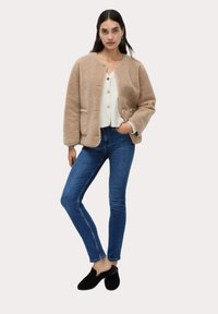 Tan fleecejacka med rund hals, guldknappdetaljer och tv å sidofickor, ihop med en vit topp, blå jeans och svarta slip-on skor.