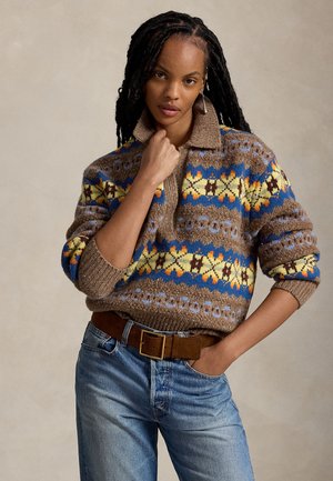 Femme aux longs cheveux tressés portant un pull en maille marron, bleu et jaune à motifs, un jean bleu et une ceinture marron sur un fond uni.