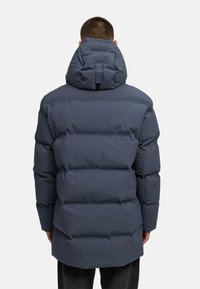 Temno modra puhovka z visokim ovratnikom, ki ima šivane predele, zadnjo kapuco in elastične manšete za tesno prileganje.