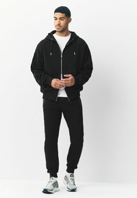 Schwarze Zip-Hoodie mit Kordelzügen, kombiniert mit schwarzen Jogginghosen. Das Model trägt ein weißes T-Shirt darunter und silberne sowie graue Sneakers.