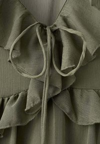 Détail d'une robe en tissu texturé vert olive avec des volants superposés et un fin nœud à nouer au niveau de l'encolure.