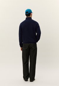 Homem de pé de costas, vestindo um boné azul, casaco azul-marinho, jeans soltos pretos e sapatos pretos, contra um fundo claro e simples.