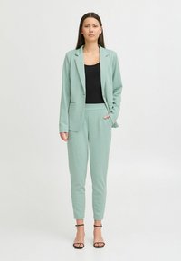 Svetlozeleni krojeni kostim, ki vključuje strukturiran blazer in zožene hlače. Usklajen z črnim topom in natikači s peto. Teksturovan material, eleganten dizajn.