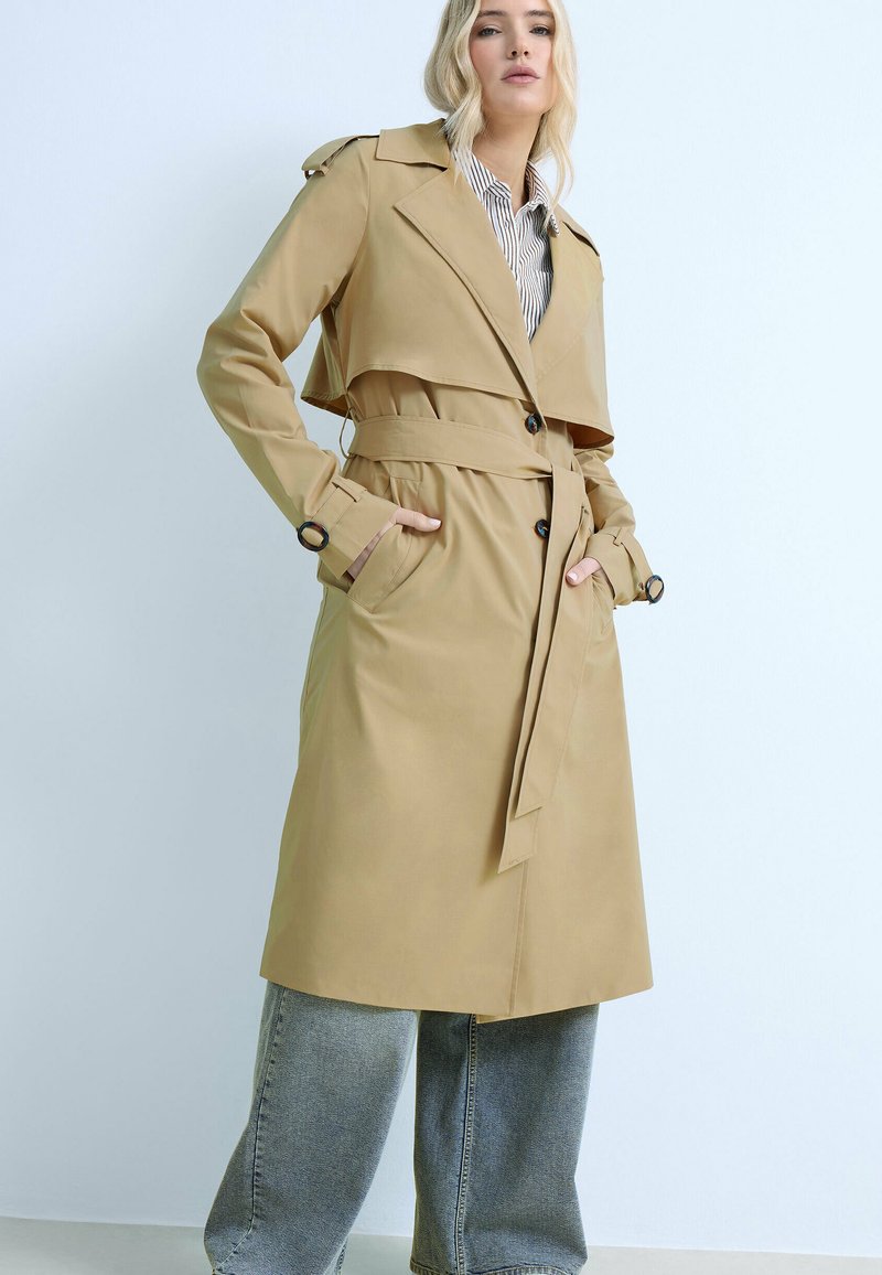 Donna bionda che indossa un trench color beige con cintura sopra una camicia a righe e jeans a gamba larga, in piedi con le mani nelle tasche del cappotto.