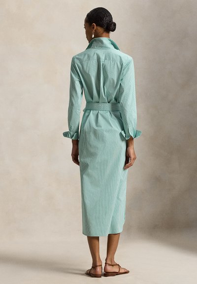 Ένα ριγέ φόρεμα-πουκάμισο σε ανοιχτό πράσινο χρώμα με γιακά, μακριά μανίκια, ζώνη στη μέση και μήκος μέχρι το μέσο της γάμπας, με μανσέτες με κουμπιά και απλό σχέδιο.
