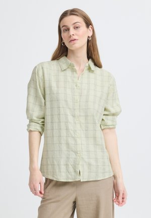 Donna che indossa una camicia a quadri verde chiaro con maniche arrotolate e pantaloni beige, in piedi davanti a uno sfondo chiaro e semplice.