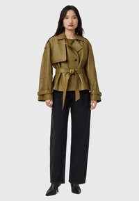 Giacca in pelle verde oliva con cintura, ampie rever, parte anteriore doppiopetto e maniche oversize, abbinata a jeans neri a gamba larga.
