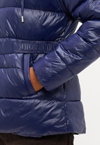 Tamsiai mėlynas pūkinis švarkas su blizgiu paviršiumi, turintis aptrauktą liemenį, elastines rankogalius ir šonines kišenes. Textūruotas siuvinėjimo raštas.