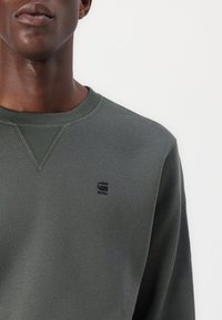 Felpa verde oliva con scollo rotondo, dettagli a coste e un piccolo logo nero sul petto a sinistra. Tessuto liscio e strutturato.