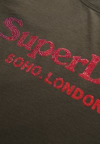 Sudadera de color verde oliva con "SuperDry" en cristales rojos en negrita, con "soho. LONDRES" impreso debajo en cristales rojos más pequeños. Textura suave.