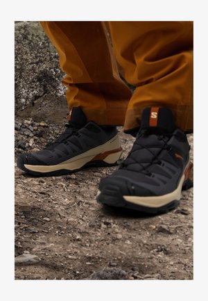 Black and tan hiking shoes with a textured surface, reinforced toe cap, and integrated lacing system, set against a rocky outdoor backdrop.