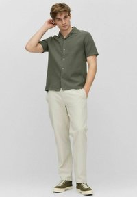 Korte mouwen, olijfgroen shirt met een gestructureerde stof, open kraag en knoopsluiting, gecombineerd met crème broek en groene schoenen.