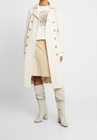 Trench coat largo de un tono beige claro con botones dorados, combinado con una blusa blanca con un diseño de logo marrón y una falda beige. Botas beige hasta la rodilla.