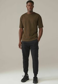 Camiseta de manga corta verde oliva combinada con pantalones cargo jogger negros que presentan bolsillos laterales y puños elásticos. Zapatillas deportivas negras completan el conjunto.