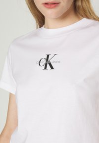 Balta medvilnės marškinėliai su juodu "CK" logotipu ir tekstu "Calvin Klein Jeans" apačioje. Trumpi rankovės ir apvalus iškirpimas. Lygus paviršius.