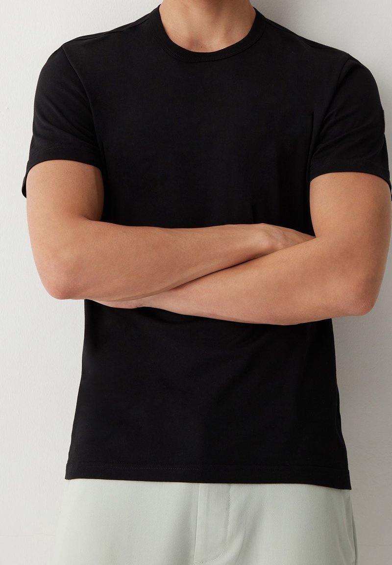 Homme les bras croisés portant un t-shirt noir uni à manches courtes et un pantalon de couleur claire sur un fond blanc.