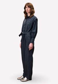 Combinaison rayée bleu marine à manches longues, avec fermoir à boutons à l'avant et ceinture à nouer. Elle comporte une poche à rabat et est portée avec des mocassins argentés.