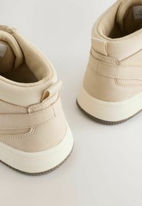 Baskets montantes beiges en matériau synthétique lisse, avec doublure en maille texturée, semelles en caoutchouc blanches et un design minimaliste.