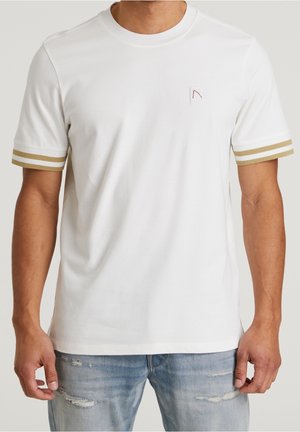 Homme portant un t-shirt blanc à manches courtes avec des doubles bandes beiges sur les manches et un petit logo sur la poitrine, associé à un jean clair déchiré.