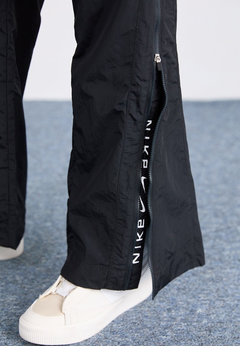 Pantalon noir zippé en matériau léger, doté de zips latéraux exposant un logo Nike blanc, associé à des baskets beiges.