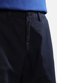 Gros plan sur la taille d'un pantalon noir avec un bouton et des passants de ceinture, porté avec une chemise bleue rentrée.