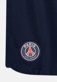 Coin de maillot de sport marine présentant le logo circulaire brodé du Paris Saint-Germain avec des accents rouges et blancs et une fleur de lys dorée.