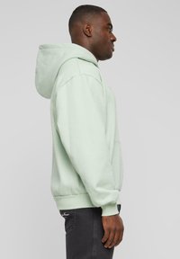 Lys grønn hettegenser med avslappet passform, med en stor hette og ribbet mansjetter. Bærer den med mørke jeans, som viser et minimalistisk design.