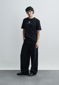 Melns kokvilnas T-krekls ar īsām piedurknēm un apaļu kakla izgriezumu, ar grafisku dizainu uz krūškurvja. Kombinēts ar platām melnām biksēm.