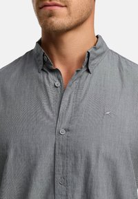 Harmaa nappipaita pehmeällä tekstuurilla, hieman rispaantuneella kauluksella ja hienovaraisella logo-ompeleella. Siinä on harmaat napit ja rento istuvuus.