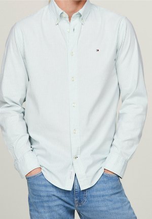 Mężczyzna w jasnoniebieskiej koszuli w paski z małym logo na piersi i niebieskich dżinsach, ręce w kieszeniach, na neutralnym tle.