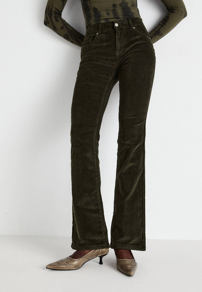 Pantaloni a zampa d'elefante in corda color verde oliva con evidenti righe verticali e tasche frontali. Abbinate a décolleté oro con punta appuntita.