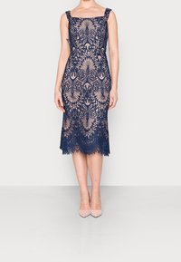 Navy blue lace dress with a square neckline, thick straps, and intricate floral patterns; knee-length with scalloped hem and nude heels.