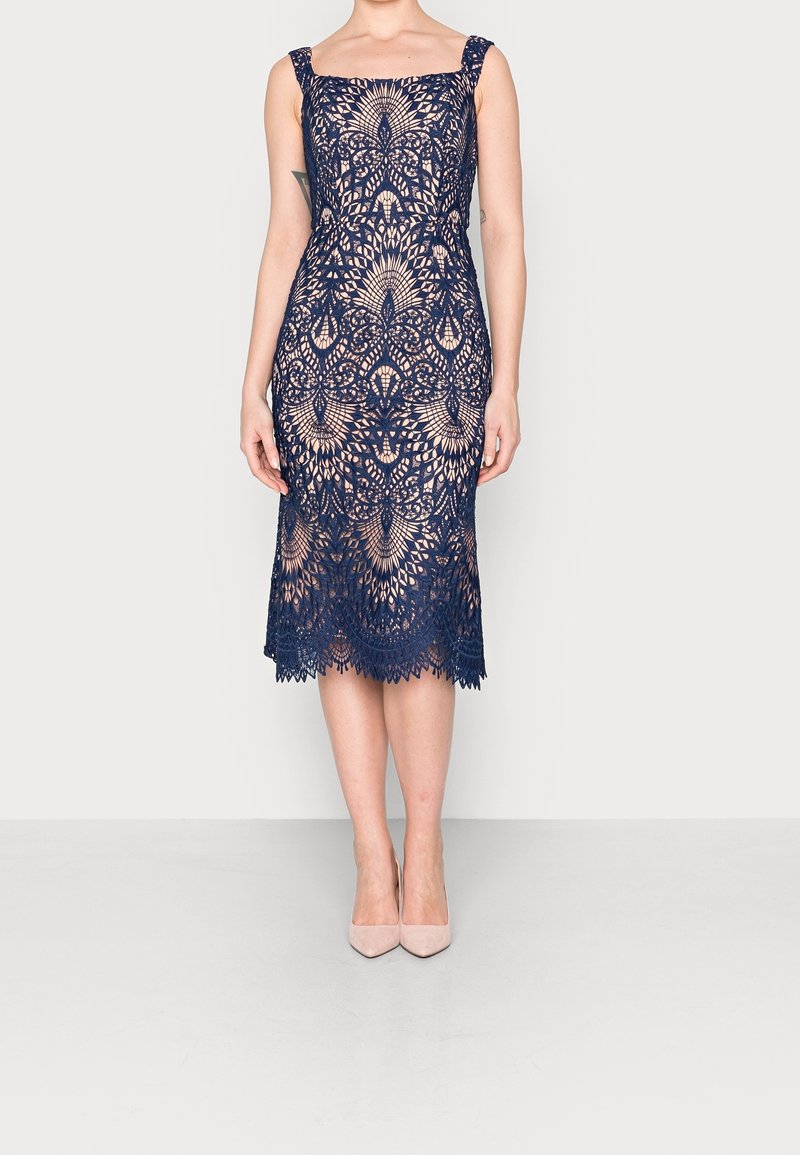 Navy blue lace dress with a square neckline, thick straps, and intricate floral patterns; knee-length with scalloped hem and nude heels.