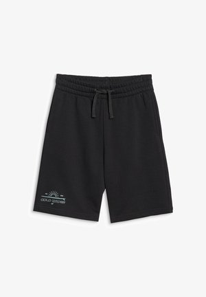 Garçon portant un sweat à capuche noir zippé et un short, un t-shirt tie-dye bleu, des chaussettes blanches et des baskets noires, debout devant un fond uni.