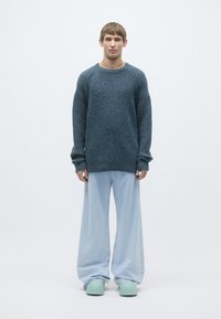 Sweater in dark blue knit with raglan sleeves, paired with loose light blue denim pants. Model wears mint green footwear. Minimalist backdrop.
