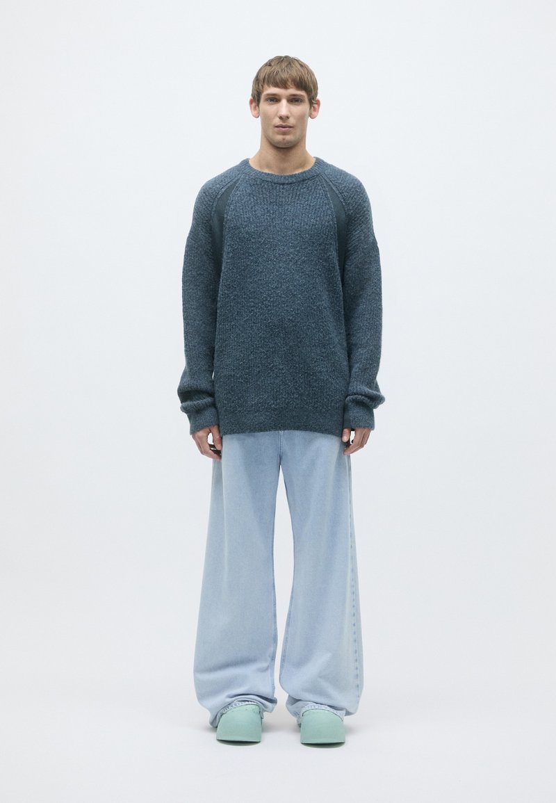 Sweater in dark blue knit with raglan sleeves, paired with loose light blue denim pants. Model wears mint green footwear. Minimalist backdrop.