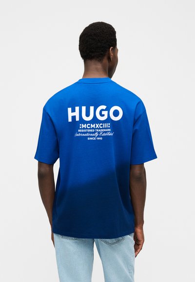 Мъж, носещ синя тениска с марка HUGO и бял надпис на гърба, както и светлосини дънки, стои пред бял едноцветен фон.