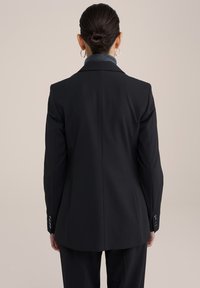 Schwarzer Blazer mit taillierter Passform, ausgestattet mit einem Schlitzausschnitt hinten, strukturierten Schultern und geknöpften Manschetten. Texturierten grauen Kragen sichtbar.
