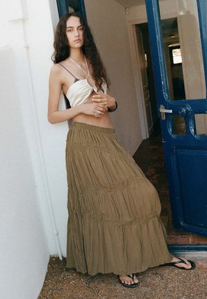 Mujer con el pelo largo que lleva una camiseta sin mangas color crema, falda larga verde oliva y sandalias negras, apoyada en una pared blanca junto a una puerta azul.