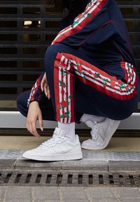 Person som sitter på huk iklädd en marinblå träningsoverall med röda blommiga ränder och vita sneakers med vita strumpor på en trottoar.