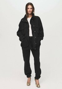 Zwarte oversized jas van textuurstof met meerdere zakken, gecombineerd met strakke zwarte broeken en hooggehakte schoenen met een patroon.