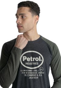 T-shirt med lange ærmer, der har grå krop og mørkegrønne ærmer. trykt logo og tekst i hvidt på forsiden. Blandingsstof af bomuld.