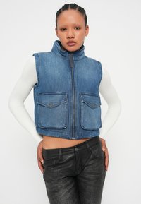 Denimvest i middels blå med høy krage, kort design, to lommer foran og glidelås foran. Teksturert stoff.
