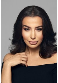 Modèle avec de longs cheveux noirs, portant un haut noir sans épaule. Peau lisse, maquillage neutre, et une main reposant sur son épaule. Fond gris.