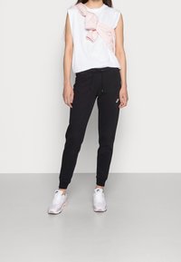 Bílý bezrukávový triko, černé zúžené joggingové kalhoty s šňůrkou a světle růžové tenisky Nike s prvky z síťoviny a gumy.
