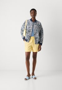 Floral bomber jacket in navy and white, light blue top, yellow shorts, and pastel blue slide sandals. The look features a crossbody bag.
