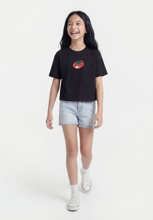 Mädchen mit langen schwarzen Haaren lächelt, geht nach vorne und trägt ein schwarzes Crop-T-Shirt mit einem roten Tomatenmotiv, helle Jeansshorts, weiße Socken und Turnschuhe.