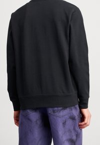 Sudadera negra de cuello redondo con un acabado texturizado, que presenta puños y dobladillo acanalados. Combinada con unos vaqueros morados, ligeramente desgastados.